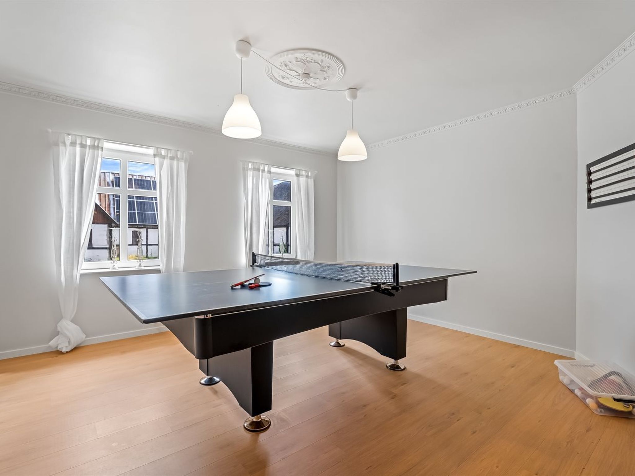 A ping pong table in a room with wooden flooring, white walls, and two windows with white curtains.