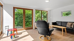Ein modernes Wohnzimmer mit einem schwarzen Ledersessel, einem Holztisch, einem rot-blauen Hocker, einer schwarzen Couch und großen Schiebeglasfenstern mit Blick auf Grünflächen.