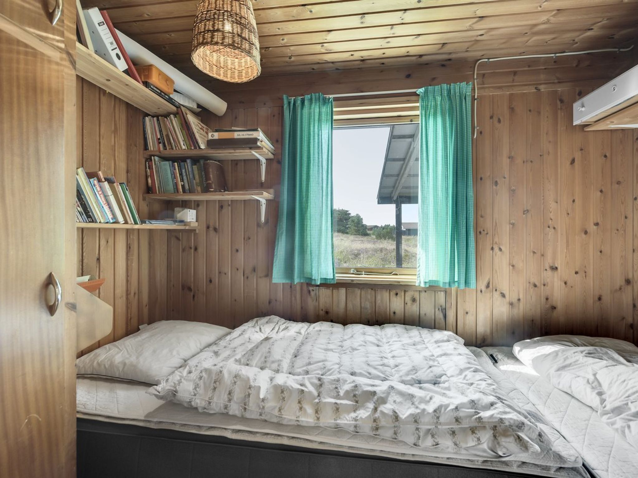 Quarto de cabana de madeira com cama, estantes e janela com cortinas.