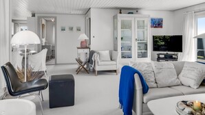 Ein modernes Wohnzimmer mit einem weißen Sofa, einem schwarzen Sessel, einem Couchtisch und einem Fernseher. Es gibt ein Schrank mit Glastüren und eine Lampe auf einem Beistelltisch.
