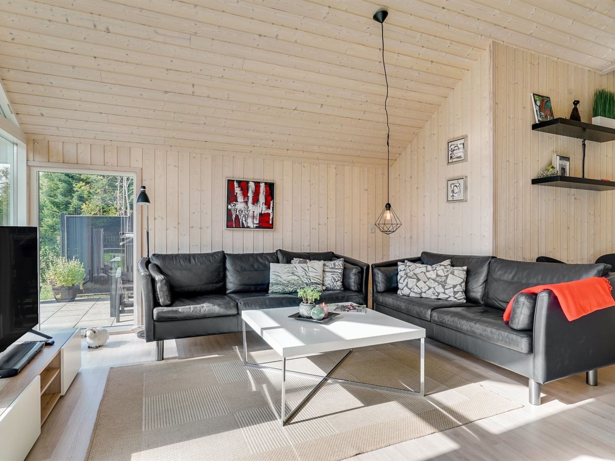 Un salon moderne avec un canapé d’angle, une table basse et un téléviseur. Le plafond est en bois et une porte donne accès à un balcon.