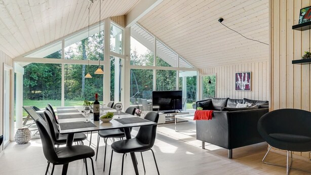 Een ruime woonkamer met een eettafel, stoelen, een zwarte bank en een televisie. Grote ramen bieden uitzicht op het omliggende landschap.