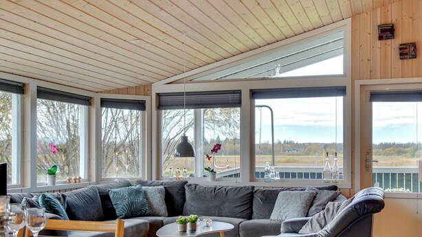Ein modernes Wohnzimmer mit einem großen Ecksofa, einem Couchtisch und einem Essbereich mit Tisch für zwei Personen. Große Fenster bieten Ausblick ins Freie.