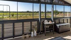 Eine überdachte Terrasse mit großen Fenstern, die einen Blick auf ein grünes Feld und Bäume bietet. Ein Tisch mit Glasplatte und eine Bank sind vorhanden.