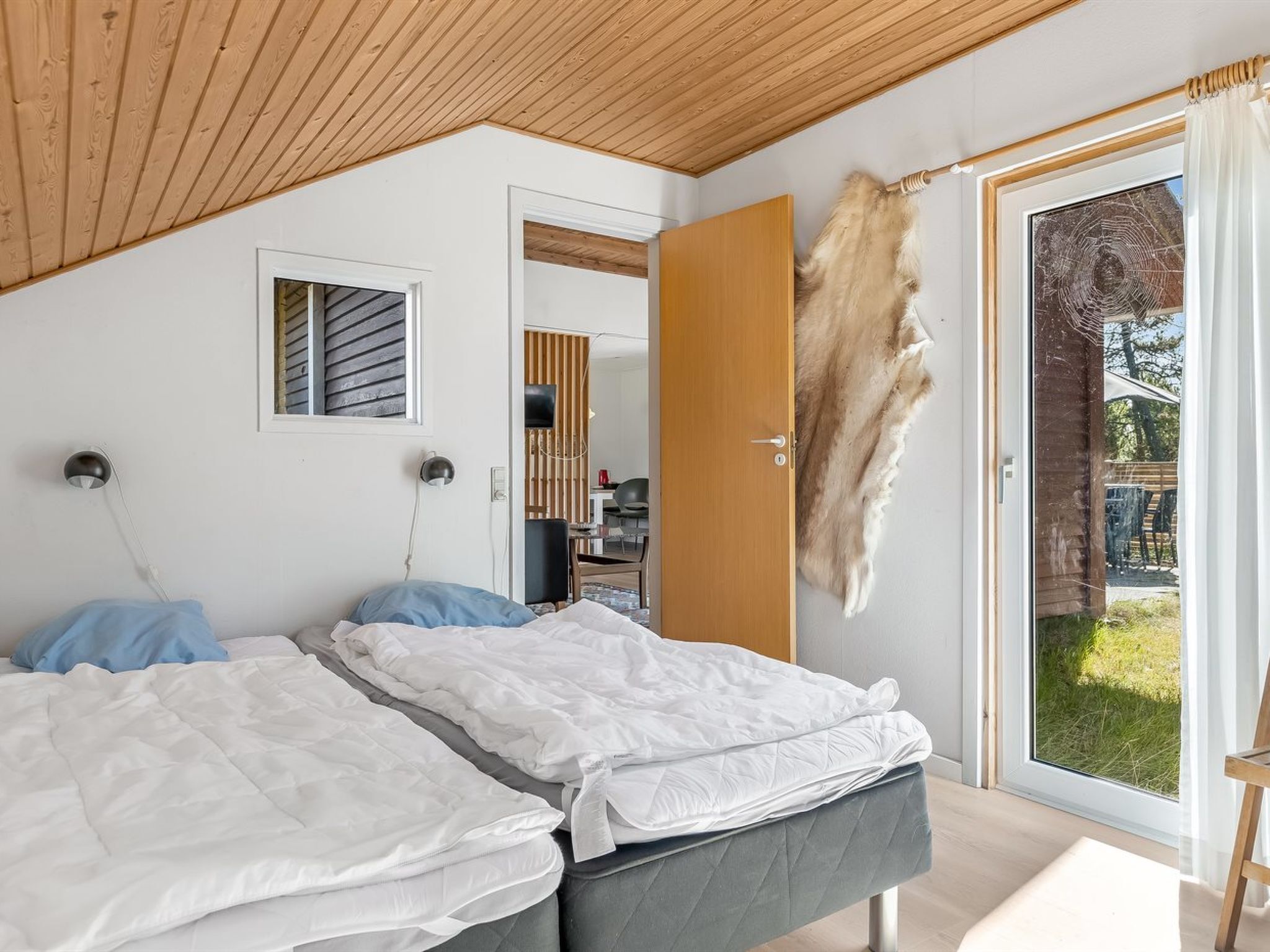 A bedroom with two beds, a wooden ceiling, a large window with a view of the outdoors, and a fur piece on the wall.