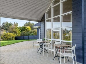 A covered patio with a table and chairs, large windows, and a view of the surrounding greenery.