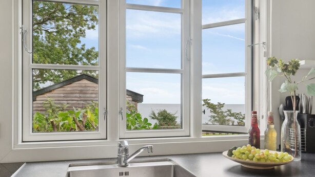 A kitchen with a stainless steel sink, a window with a view of a wooden house and greenery, and a bowl of grapes on the counter. - "Alfred" - 25m from the sea by Interhome (Gudhjem)