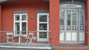 Ein Gebäude mit roter Fassade, weiß gefassten Fenstern, einem Balkon mit Gitter und Sitzgelegenheiten sowie einer Treppe, die zu einer Tür mit Glasfeldern führt.