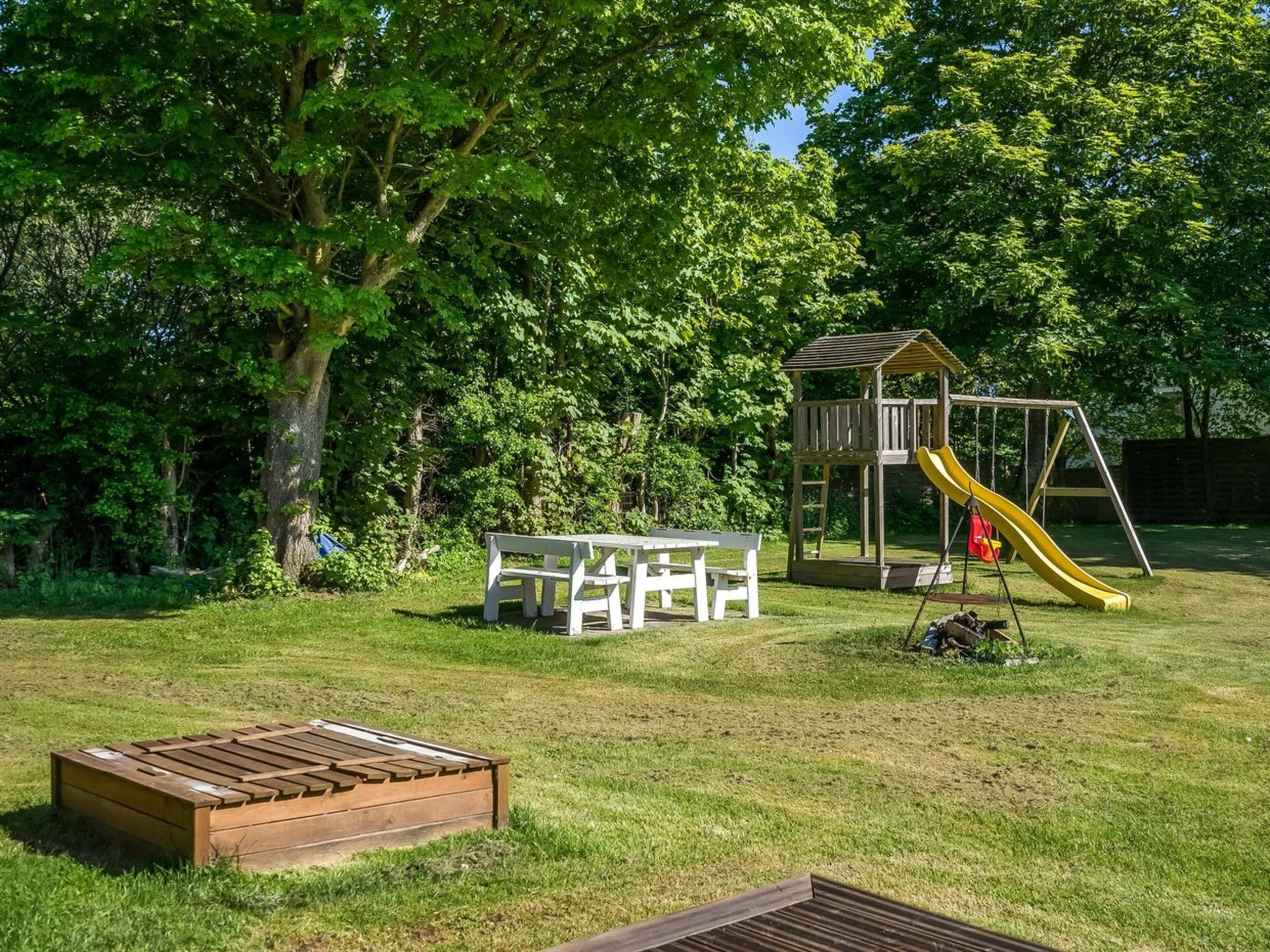 Un jardin avec une cabane en bois, un toboggan jaune, un banc blanc et une table en bois.