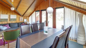 Een ruime kamer met een hoge plafond, houten balken, een eettafel met stoelen, een bank en een groot raam met gordijnen.