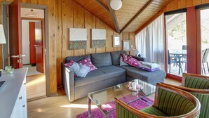 Een gezellig chaletinterieur met een houten plafond, een grijze hoekbank, een glazen salontafel en een groene fauteuil.