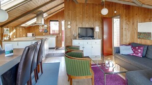 Een gezellige, goed verlichte kamer met een hoge plafond, houten wanden, een eethoek met stoelen en een tafel, een bank en een televisie.
