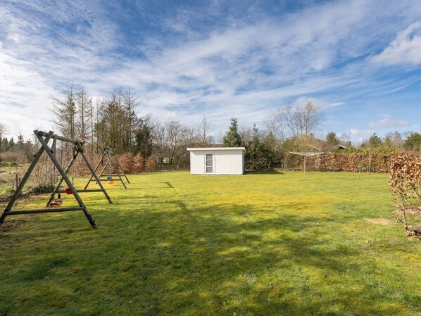 Un patio trasero con césped, un conjunto de columpios, un cobertizo pequeño y una pérgola.