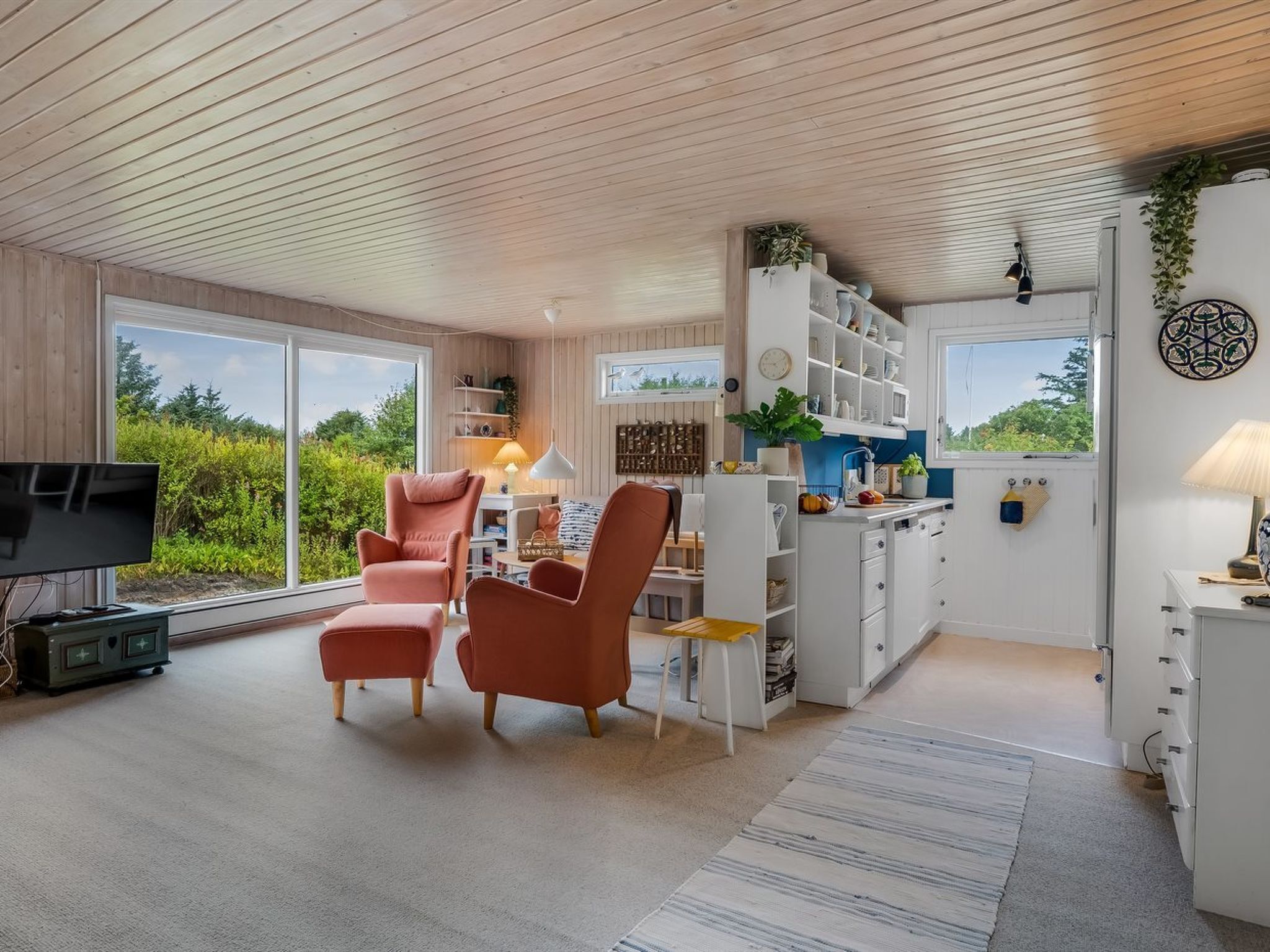 A modern living room with a television, two armchairs, a coffee table, and a kitchen area with white cabinets and a window.