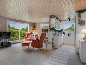 A modern living room with a television, two armchairs, a coffee table, and a kitchen area with white cabinets and a window.