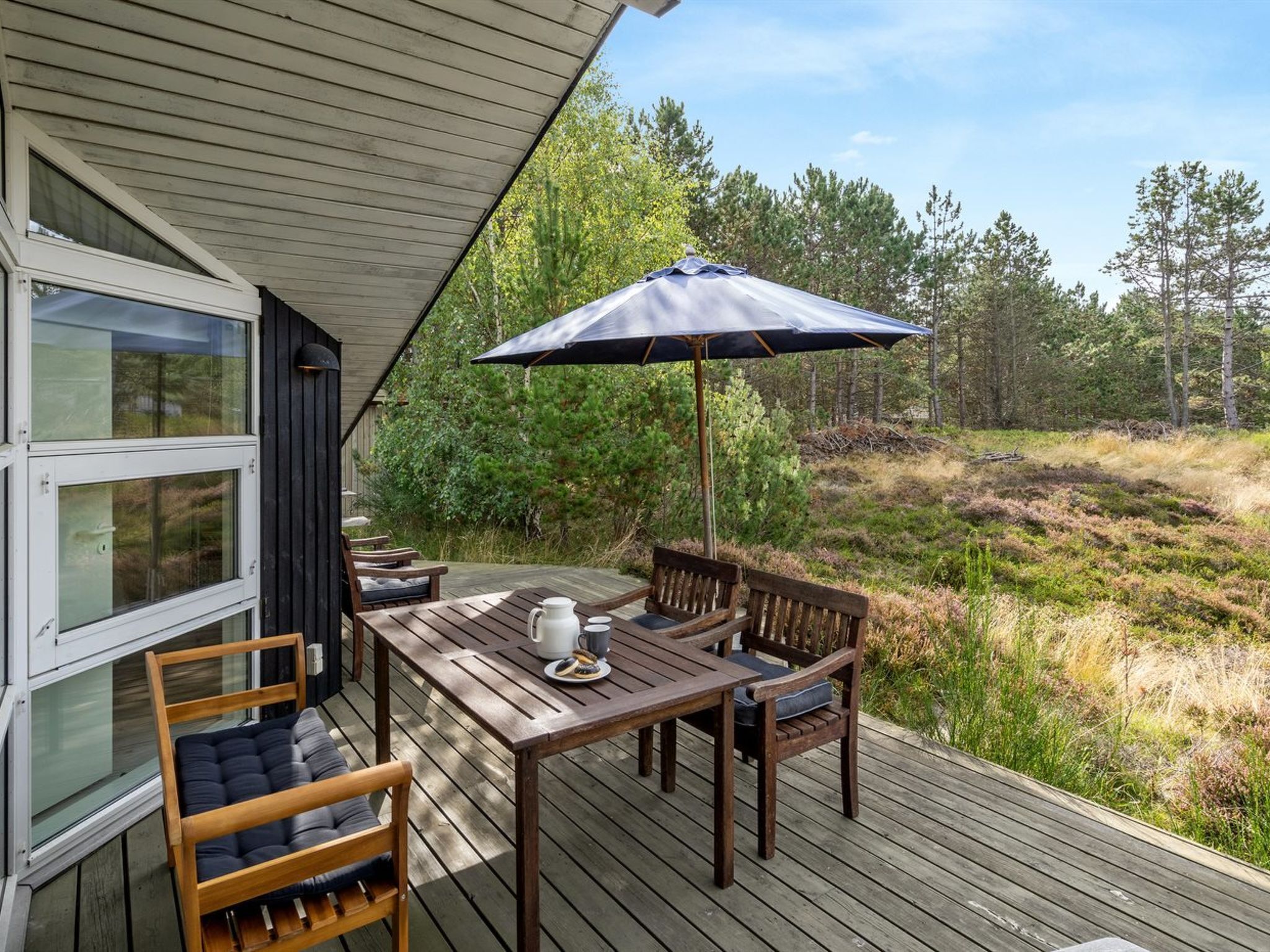 A wooden deck with a table and chairs, a large umbrella, and a view of a forested area.