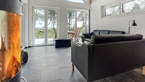 Ein großzügiger Wohnbereich mit einem Kamin, einem dunkelblauen Ledersofa und Blick ins Freie durch große Fenster.