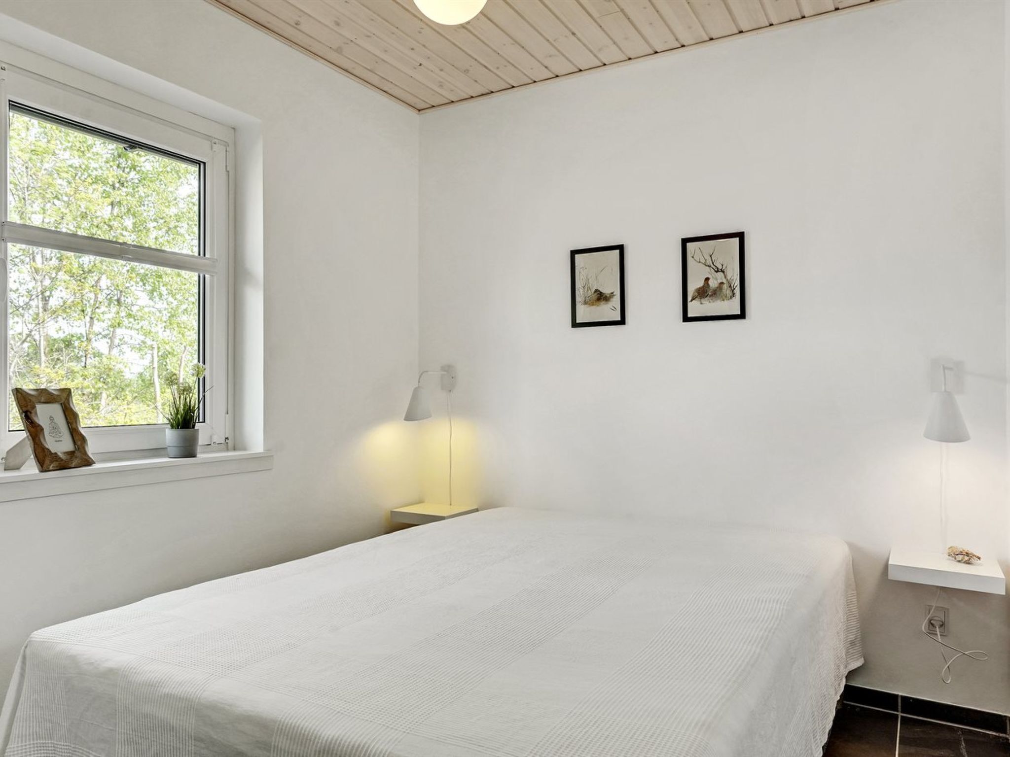 Ein Schlafzimmer mit großem Fenster, einem Bett mit weißer Bettdecke, zwei gerahmten Bildern an der Wand, einem Nachttisch mit Lampe und einem kleinen Regal mit Pflanze.