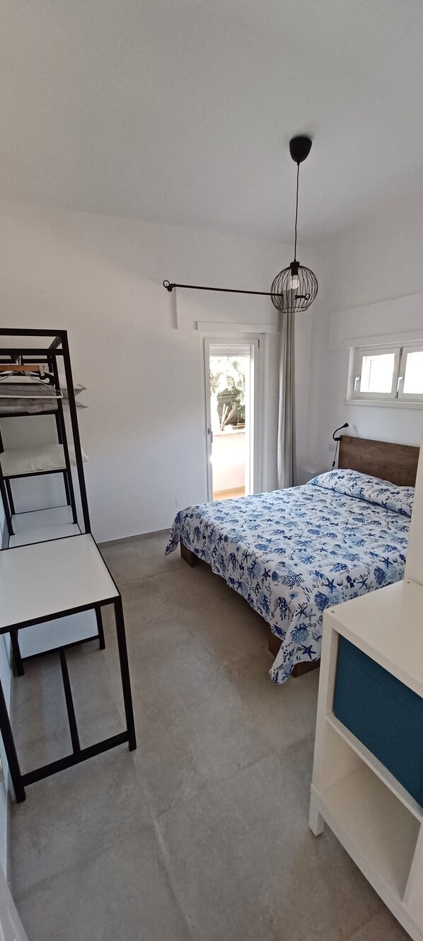 A bedroom with a bed, a desk, a hanging light fixture, and a view through a door.