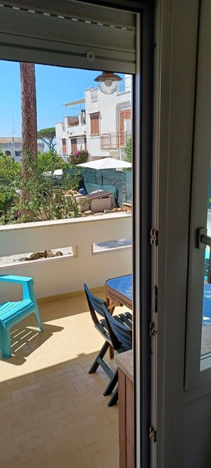 A balcony with a table and chairs, a blue plastic chair, and a view of buildings and a tree.