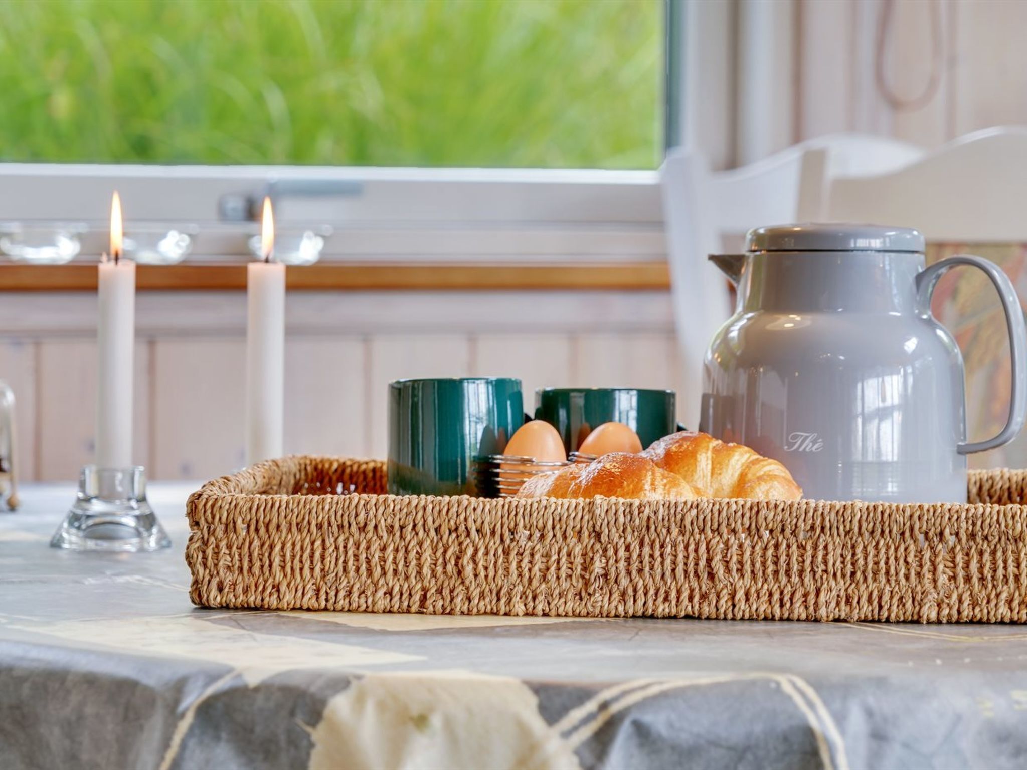 A breakfast setup with croissants, eggs, and a coffee pot on a table with candles and a view of greenery.
