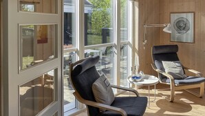 Una habitación moderna con un gran ventanal corredero que da a un espacio exterior, dos sillones tapizados, una mesita redonda y una lámpara de pie.