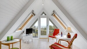Una habitación con techos altos y techo inclinado, un televisor sobre una mesa blanca, una silla roja, una lámpara y vistas al exterior.