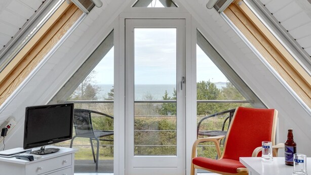 Una habitación con techo inclinado, un amplio ventanal corredero, un escritorio con computadora, una silla roja y una mesa con una botella y vasos.
