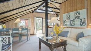 A cozy room with a wooden ceiling, a dining area with chairs and a table, a sofa with cushions, and a coffee table with flowers.