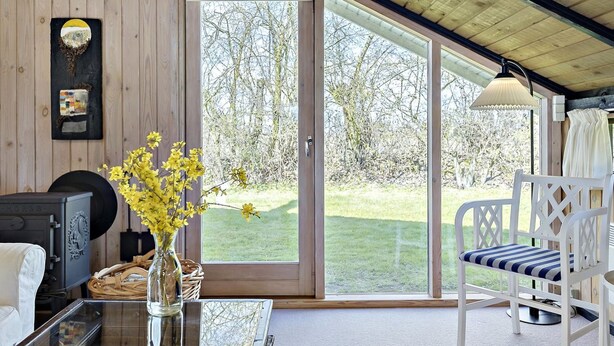 A room with a glass door leading to a garden, a white chair, a table with a vase of flowers, and a fireplace.