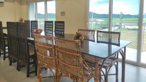 A dining area with a glass table and wicker chairs, large windows with a view of a green landscape. - Mayur Gardens (Sonapur)