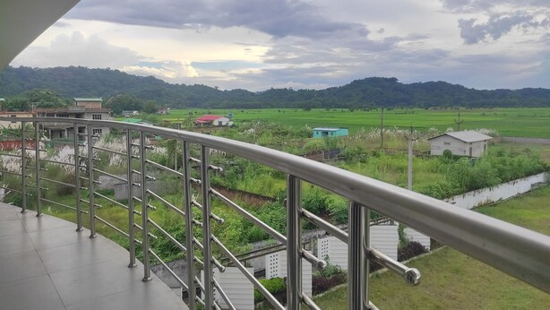 A balcony with a metal railing overlooking a green landscape with fields and a few buildings. - Mayur Gardens (Sonapur)