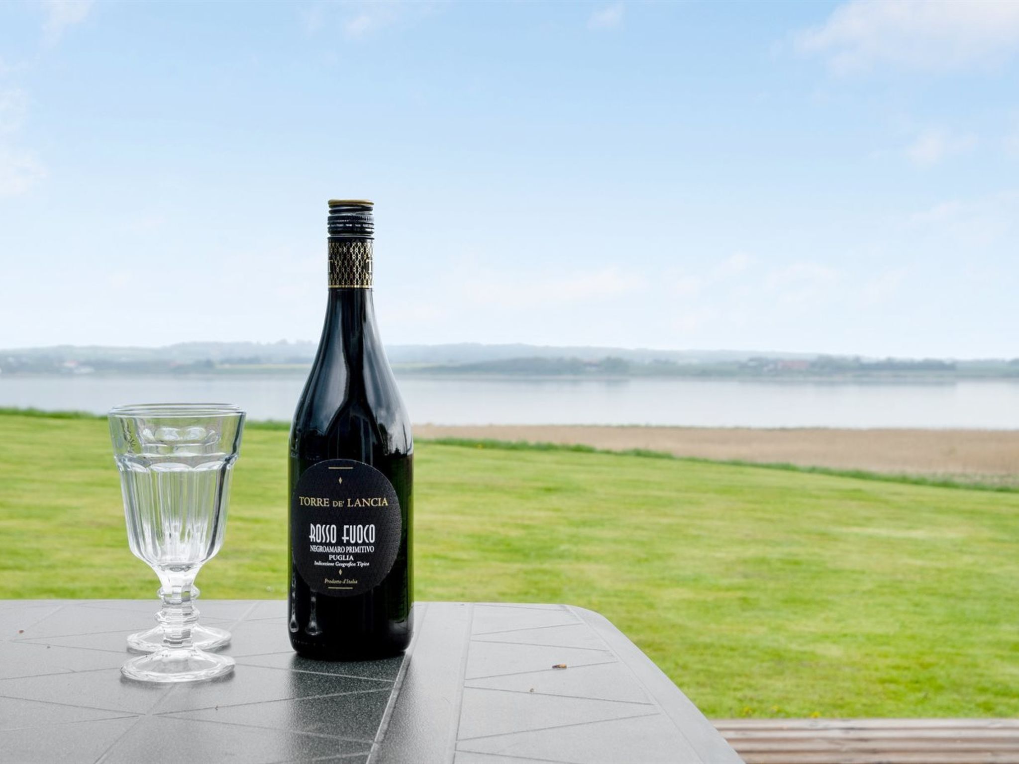 Una botella de vino rosso fuoco y una copa sobre una mesa con vistas a un lago y colinas distantes.