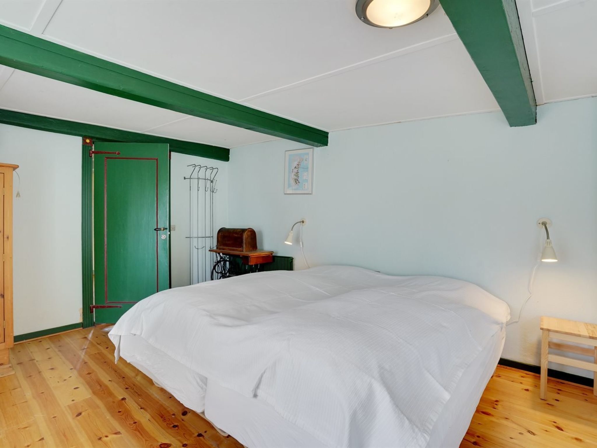 Een slaapkamer met een groot bed, een houten vloer, een groene deur, een kapstok en twee bedlampjes.