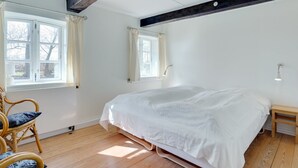 Een slaapkamer met een groot bed, houten vloer en zichtbare houten balken. Er zijn twee ramen met gordijnen, een stoel en een nachtkastje.