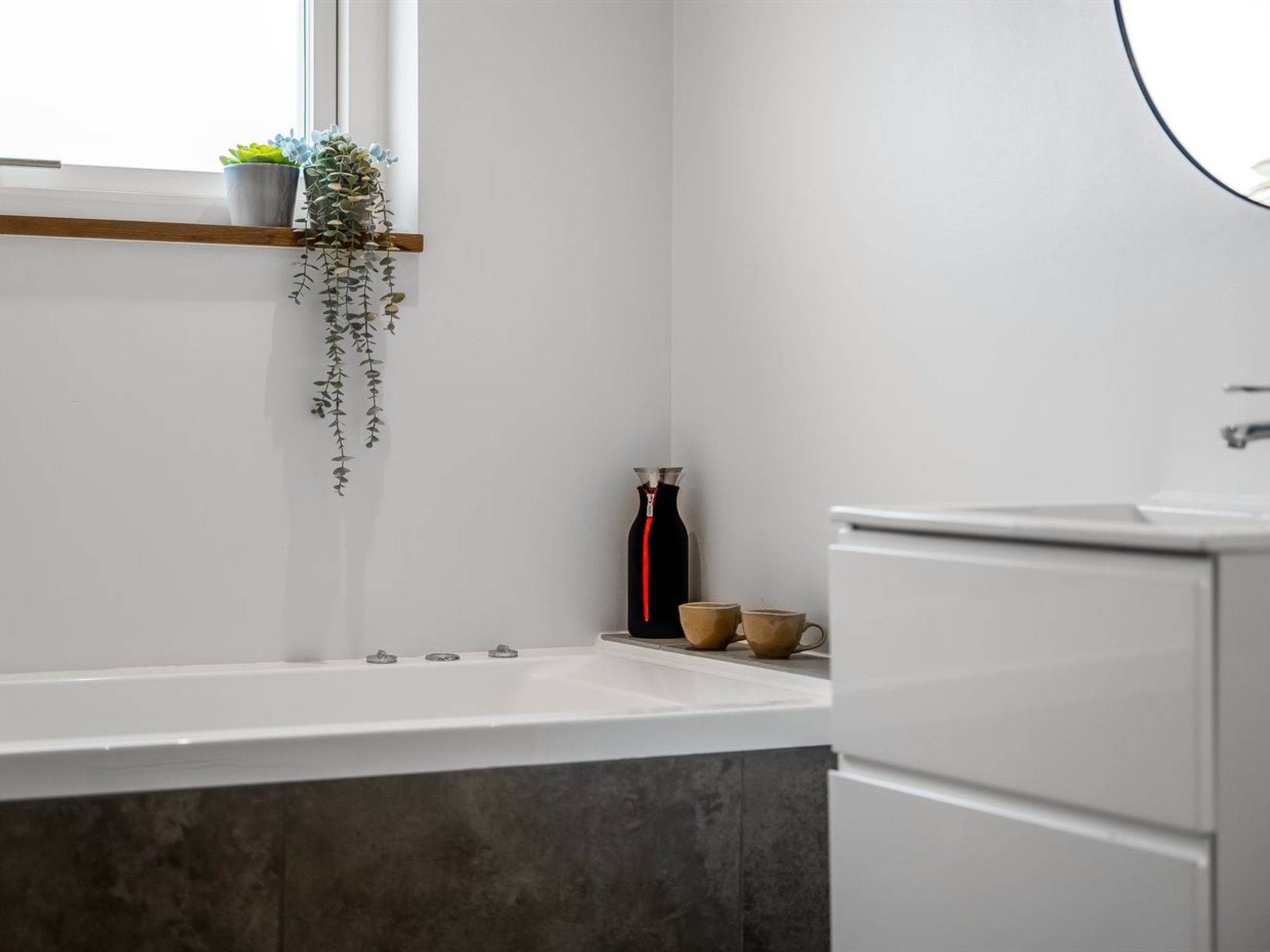 A bathroom with a bathtub, a window with plants, and a cabinet.