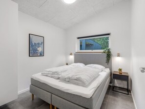 Une chambre moderne avec un lit gris, du linge de lit blanc, une œuvre d’art encadrée, une table de chevet avec une lampe et une fenêtre donnant sur un paysage verdoyant.