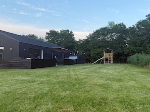 A modern building with a flat roof, a covered outdoor seating area, and a wooden play structure in a grassy yard.