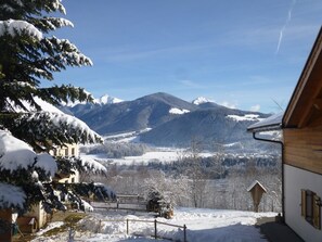 Un paysage enneigé avec des montagnes en arrière-plan, un bâtiment en bois doté d’un toit en pente et une cour clôturée.