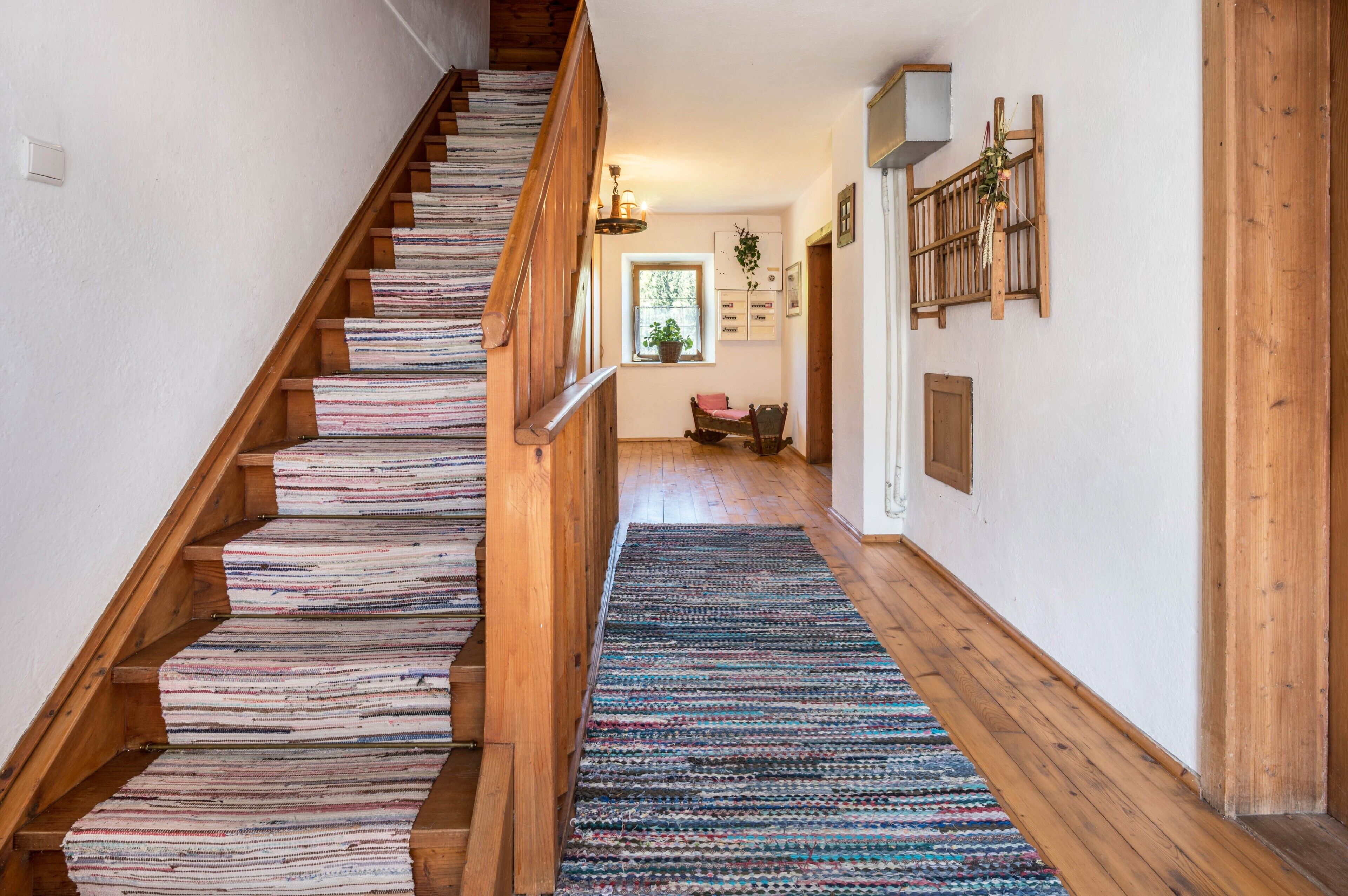 A wooden staircase with a patterned rug leading to a room with a window, a chair, and a wall-mounted shelf.