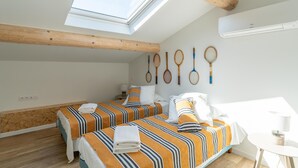 Una habitación con dos camas, un tragaluz, vigas de madera, un equipo de aire acondicionado y raquetas de tenis en la pared.
