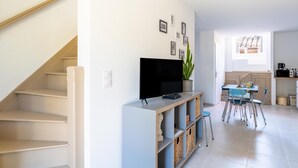 Una escalera conduce a una sala de estar con televisor, una mesa de comedor con sillas y una zona de cocina con ventana.
