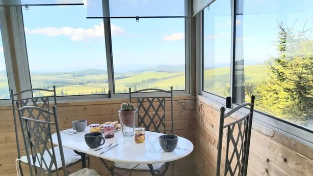 Una terraza de madera con una mesa, sillas, una planta en maceta y vistas a colinas ondulantes.