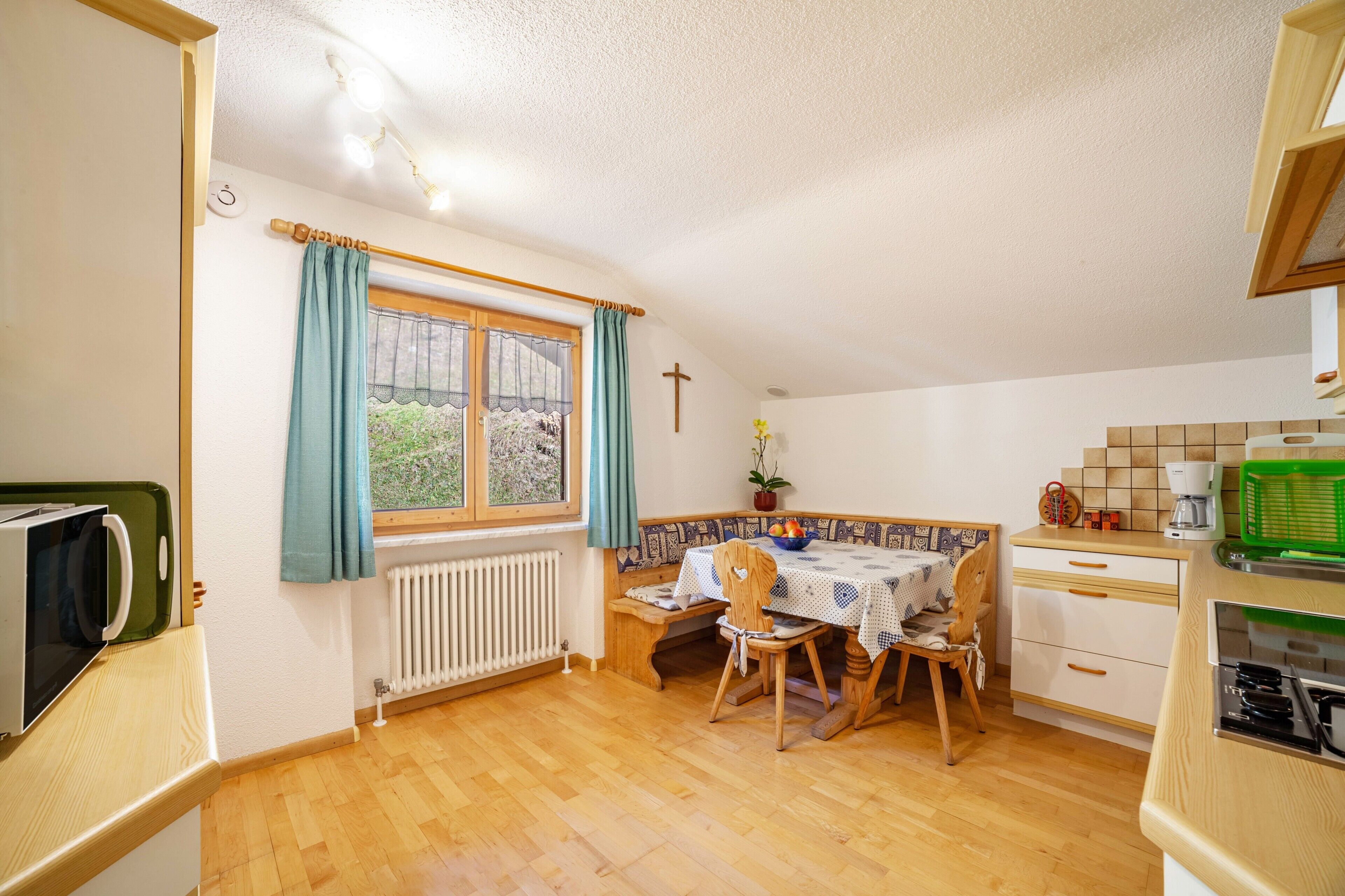 Een gezellige keuken met een eettafel, houten stoelen, een radiator, een raam met gordijnen en een kruis aan de muur.