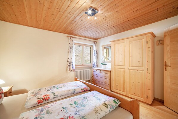 Un dormitorio con techo de madera, dos camas con ropa de cama floral, un armario de madera y una ventana con cortinas.