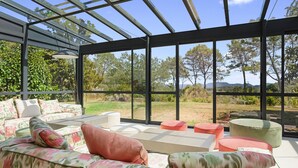 Ein Wintergarten mit einer Couch, Poufs und großen Fenstern mit Blick auf Grünflächen.