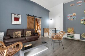 Un salon moderne avec un canapé en cuir marron, des fauteuils en osier, une console blanche et un mur décoré sur le thème du voyage.