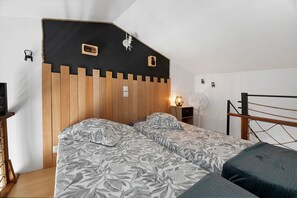 Een slaapkamer met een schuine kap, een houten hoofdbord, twee bedden met beddengoed met een patroon, een nachtkastje met een lamp en een kleine ventilator.