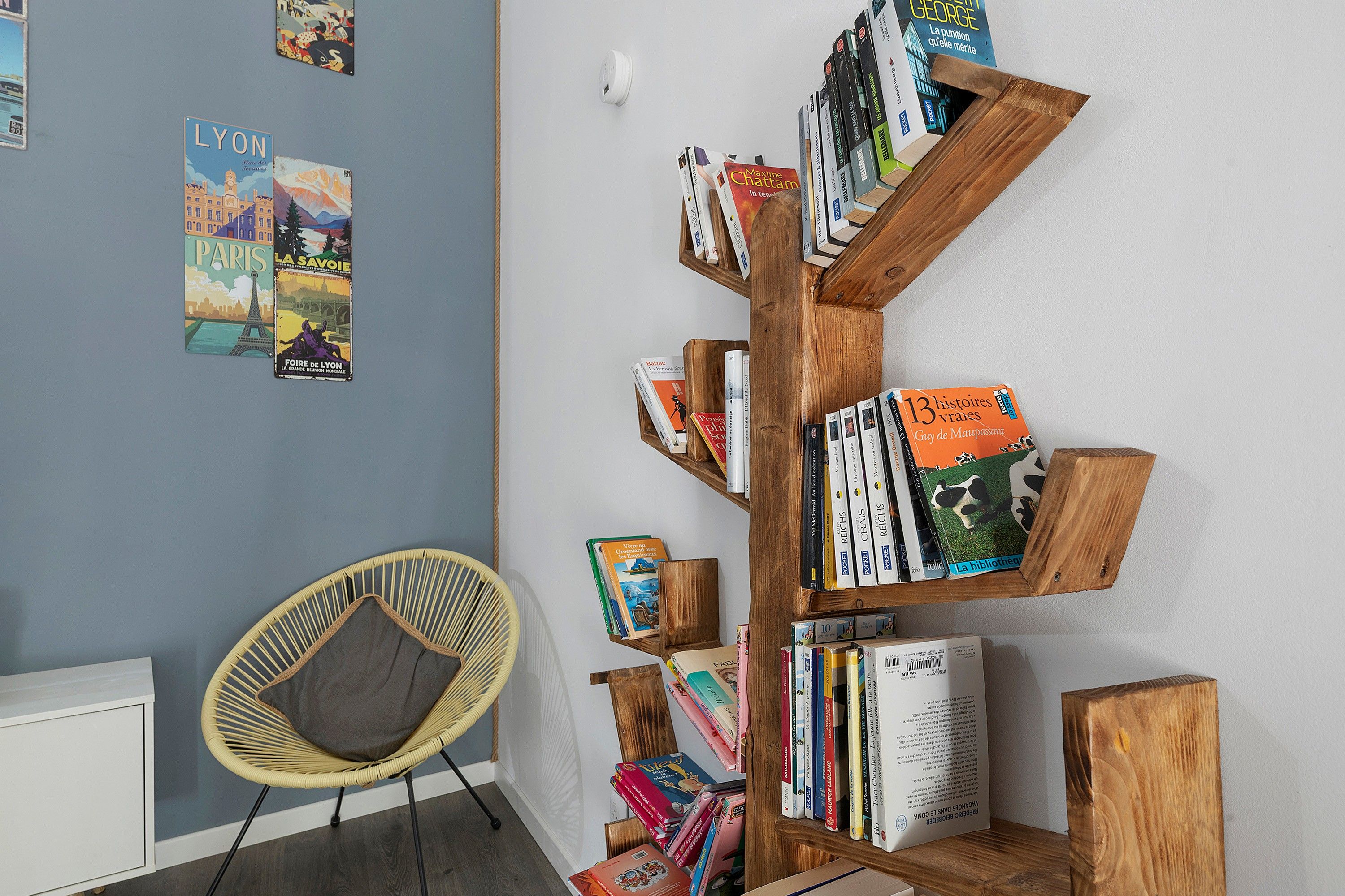 Una habitació amb una estanteria de fusta plena de llibres, una cadira de vimet i una paret decorada amb cartells de viatges.