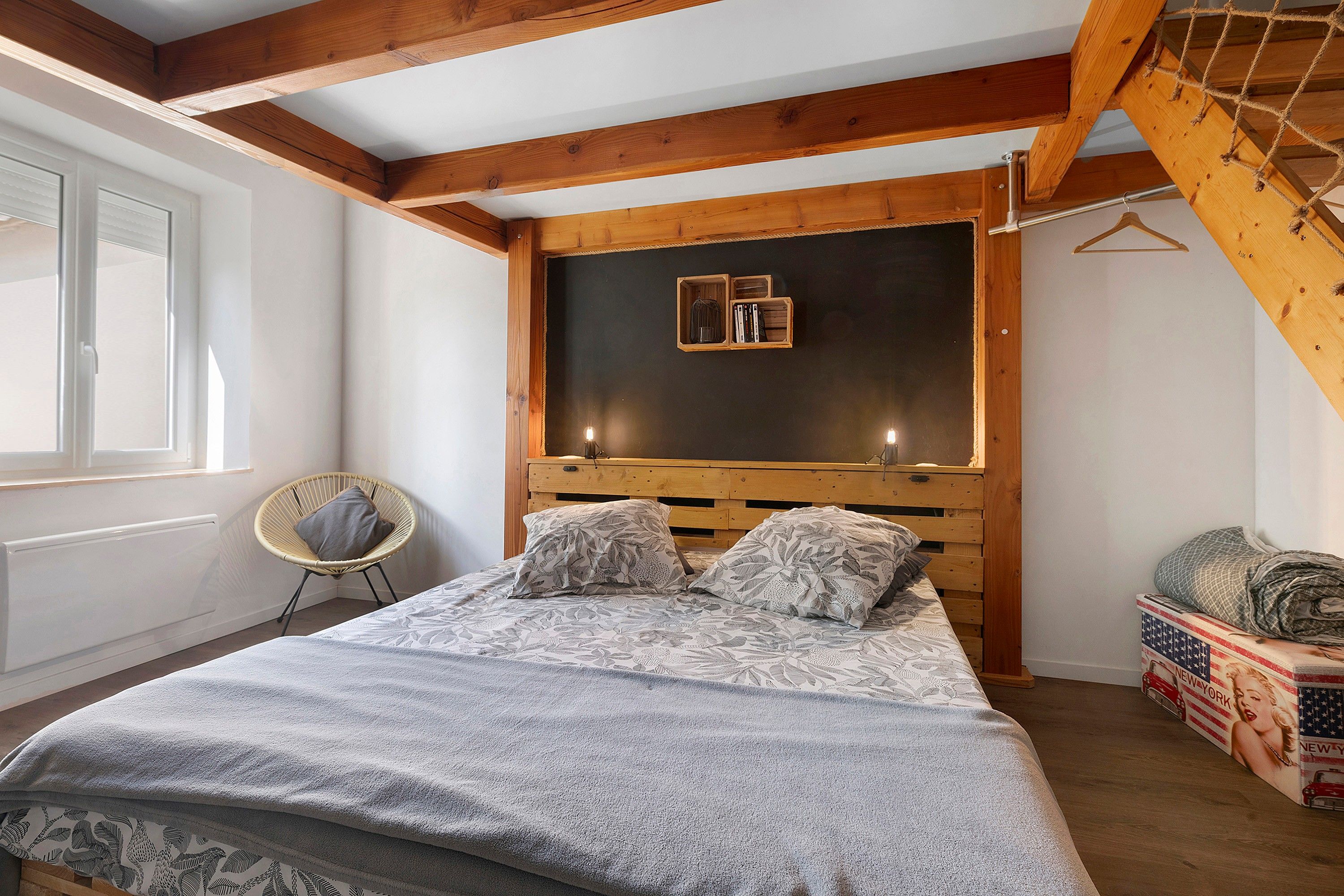 A bedroom with a wooden headboard, a bed with patterned bedding, a chair, and a small shelf with books.